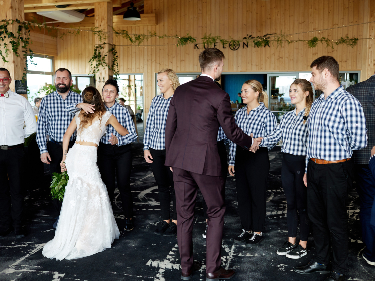 Para młoda witająca się z zespołem obsługi w szachownicowych koszulach w środku drewnianej restauracji, przygotowanej na przyjęcie weselne