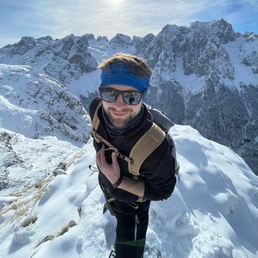 Mężczyzna w zimowym stroju, z plecakiem, uśmiecha się do kamery, robiąc selfie z przepięknym, śnieżnym górskim krajobrazem w tle, nosząc niebieską opaskę i okulary przeciwsłoneczne